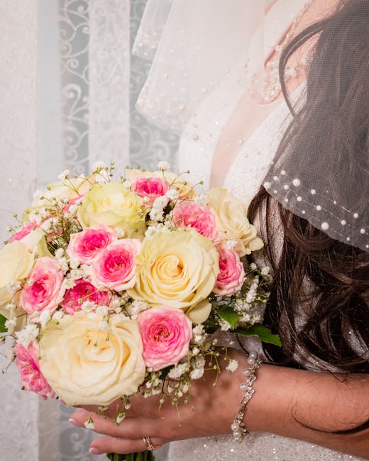 A bride holding a bouquet of fresh flowers consisting of creamy white roses and vibrant pink roses, complemented by delicate white baby's breath. The bouquet is arranged in a rounded, lush style, wrap