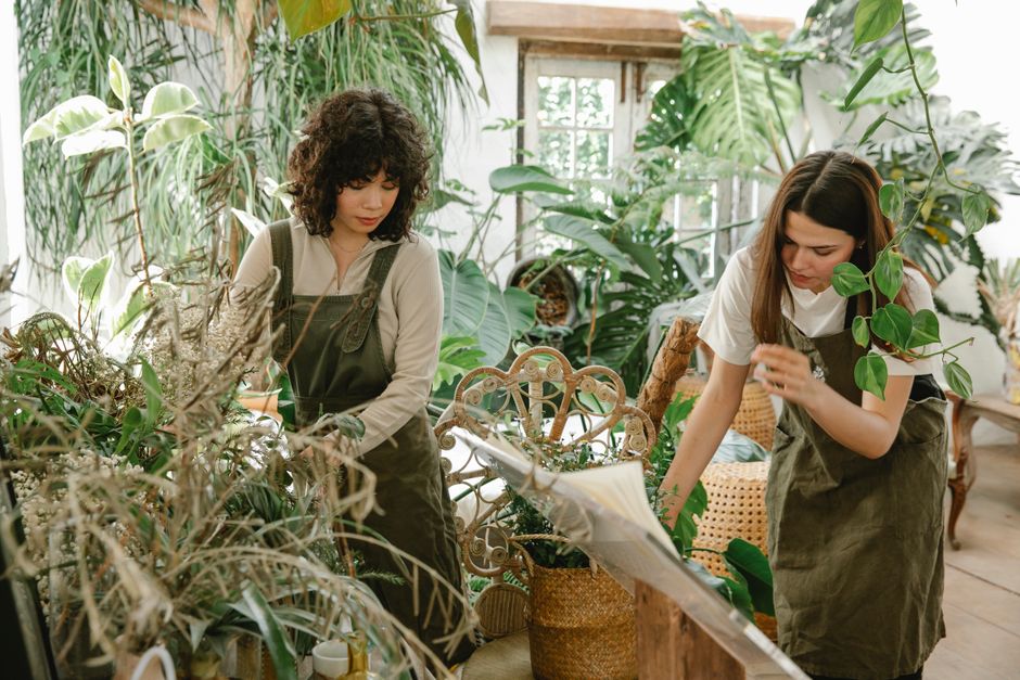 Two women working together in a lush florist studio filled with various green plants and leaves. The woman on the left has curly dark hair, wearing a beige top with olive green overalls, carefully arr