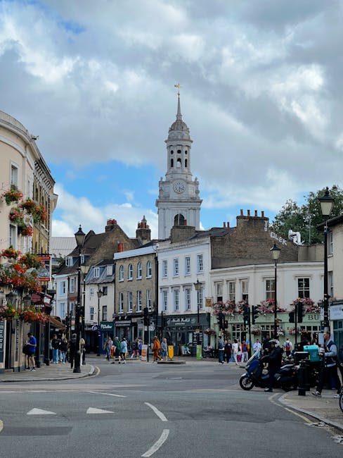 A vibrant street scene in West Hampstead featuring a floral bouquet held by a person in the foreground. The bouquet includes a mix of fresh flowers in shades of pink, purple, and white, accented with